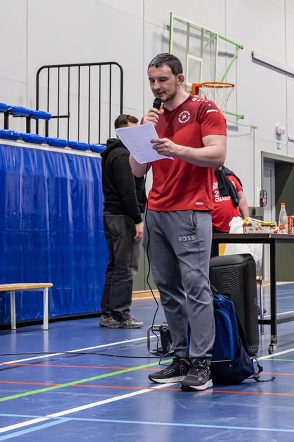 Muscular man in red sports shirt reads from paper while speaking into microphone in indoor sports hall.
