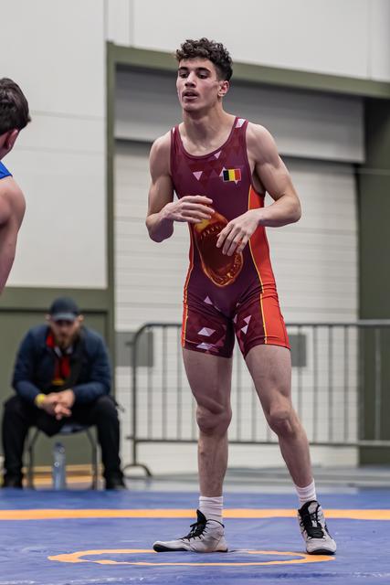 Young Belgian wrestler in maroon singlet stands alert on blue mat, hands ready, focused expression during bout.