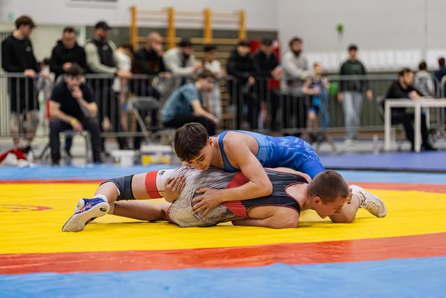 Wrestler in blue singlet pins opponent to yellow mat, showing focused determination during indoor tournament.