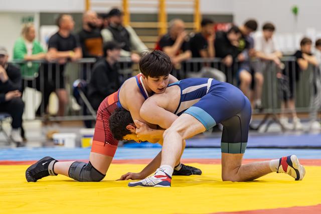 Wrestler in blue singlet dominates opponent in red, both on knees grappling intensely on yellow mat, crowd watching.