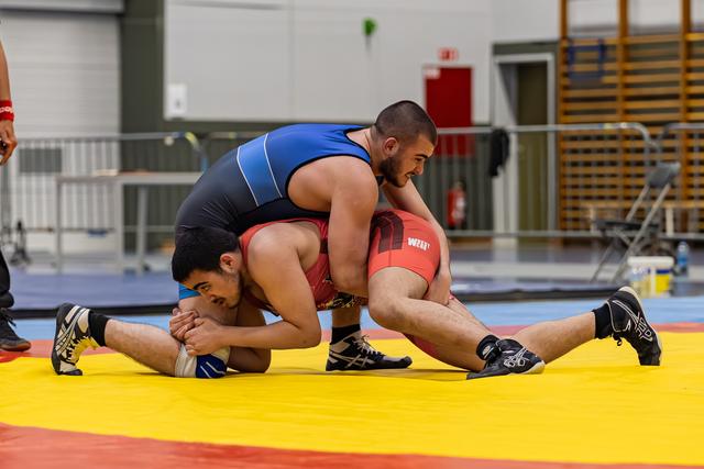 Wrestler in blue singlet controls opponent in red, pinning him down on a yellow and red mat indoors.