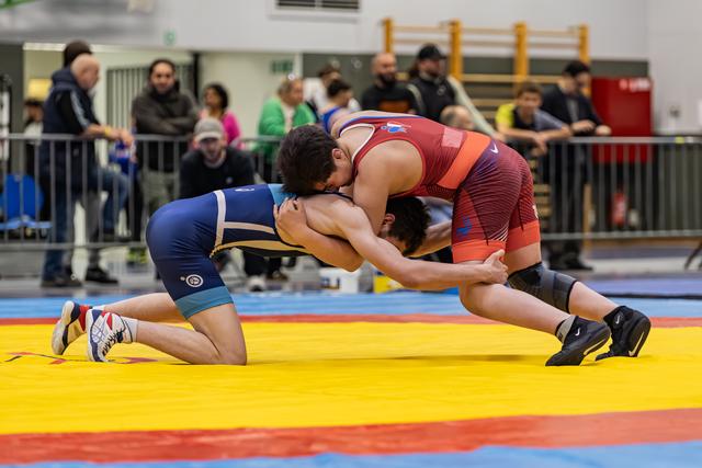 Two wrestlers lock heads in a tight clinch on a yellow mat, blue singlet athlete on one knee fighting for control.