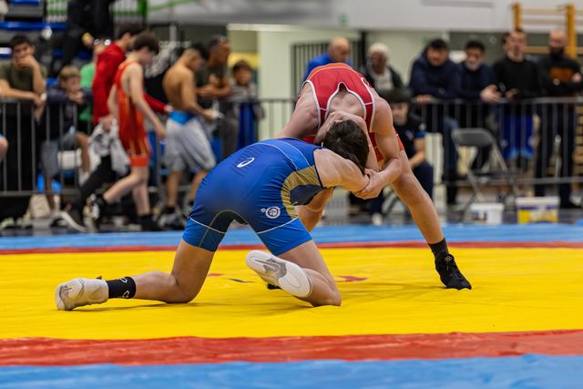 Wrestler in blue singlet on knees, locked in tight clinch with red-clad opponent on colorful mat, intense struggle.