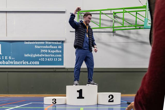 Young male athlete raises fist in triumph on 1st place podium, wearing a gold medal, smiling in an indoor sports hall.