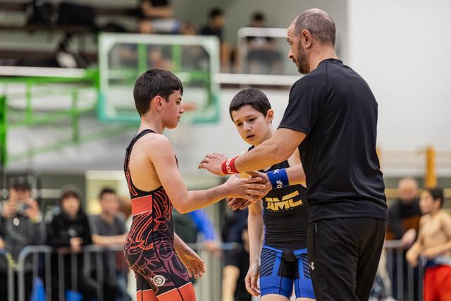 Adult coach facilitates handshake between two young wrestlers in singlets, overseeing sportsmanship on the mat.