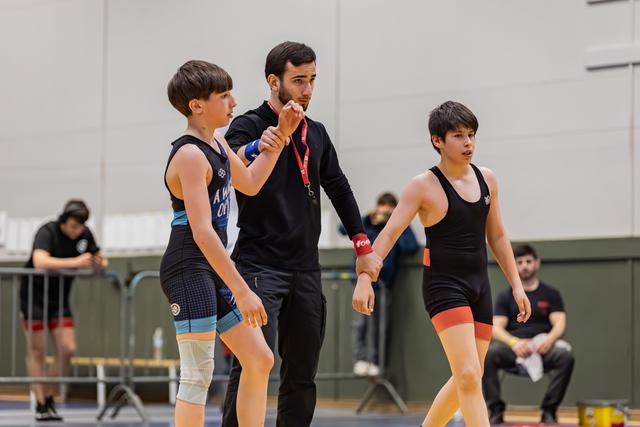 A coach in black gives focused instruction to two young wrestlers in a gymnasium, one in blue, one in black singlet.