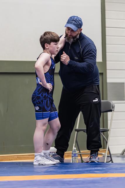 Bearded coach in navy cap leans in to give focused advice to a young wrestler in a blue singlet on the mat.