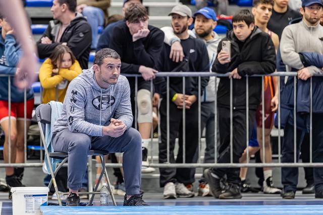 Coach in Under Armour hoodie sits intently at ringside, hands clasped, watching the match with focused concentration.