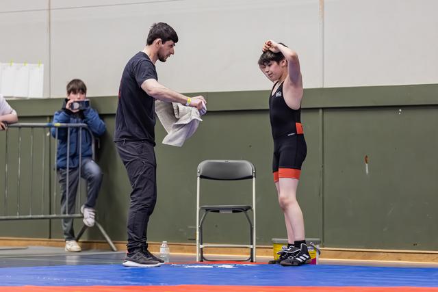 Coach hands a towel to a young wrestler in a black singlet, who stretches arm overhead, resting between bouts on the mat.
