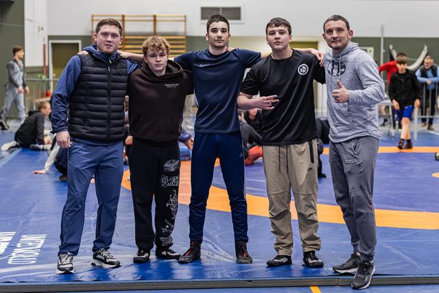 Five young wrestlers pose arm-in-arm on the blue mat, smiling confidently at a training hall event.