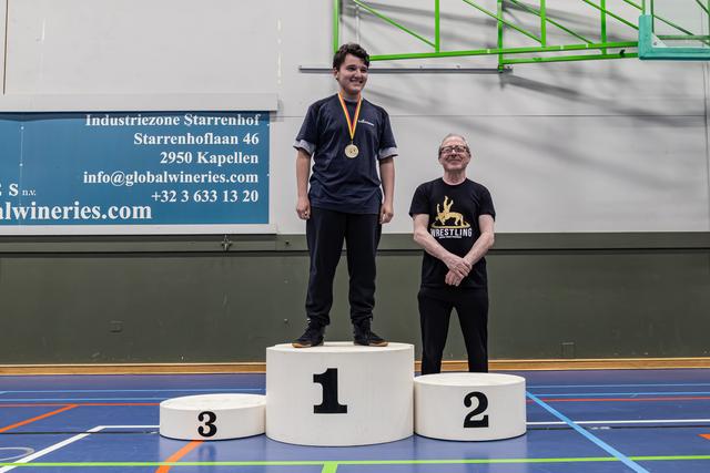 Young man smiling proudly on 1st place podium wearing gold medal, older man standing beside 2nd place podium in wrestling shirt.