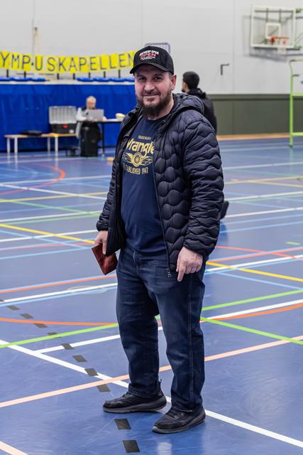Bearded man in Wrangler tee and black jacket stands relaxed on a multi-sport gymnasium floor, smiling at camera.