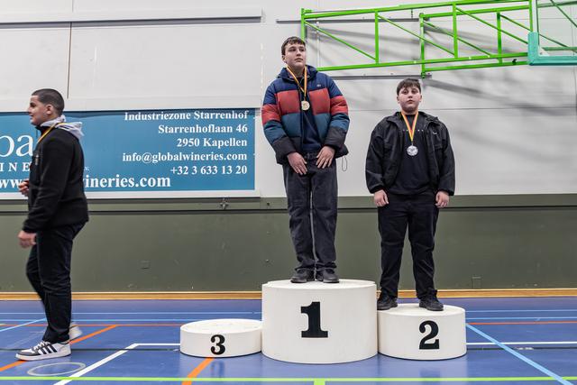 Two boys stand on podium positions 1 and 2 wearing medals, while a third walks past position 3 in an indoor sports hall.