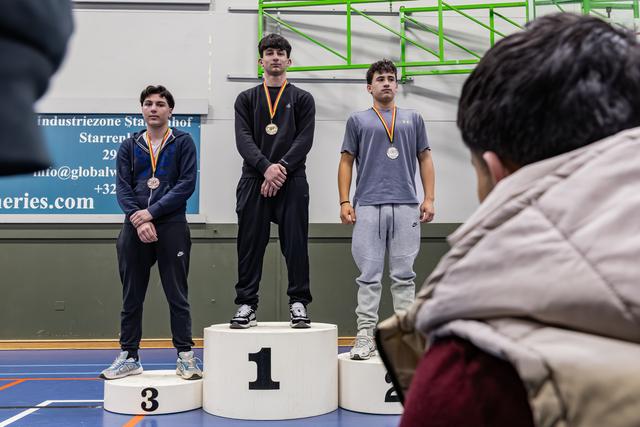Three young male athletes stand on podium positions 1, 2, and 3, wearing medals, looking solemn in a gymnasium.