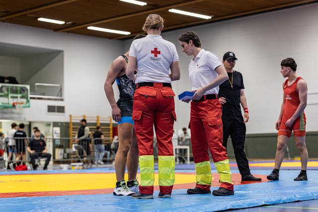 Two Red Cross paramedics consult with a wrestler on the mat during a break, while a referee and athlete look on.