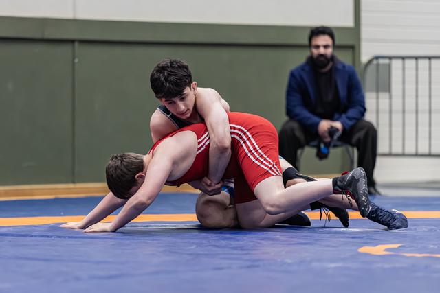Young wrestler in red dominates opponent on blue mat, controlling from above while coach watches from background.