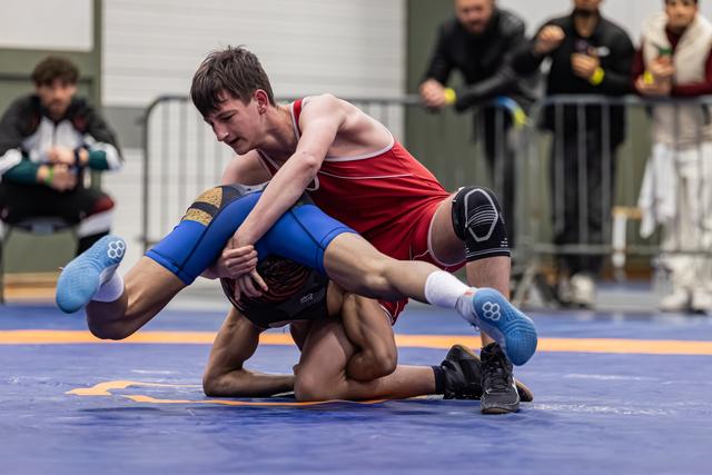 Young wrestler in red singlet drives opponent to the mat with intense focus during a competitive match.
