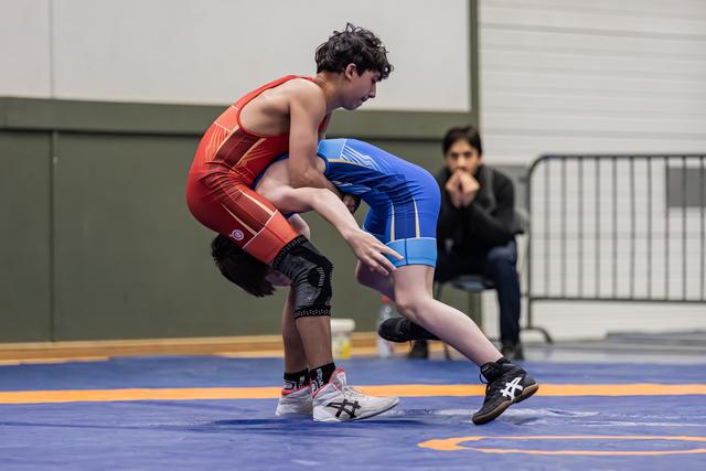 Young wrestler in red drives opponent in blue backward across the mat, both athletes focused and low in a grappling stance.