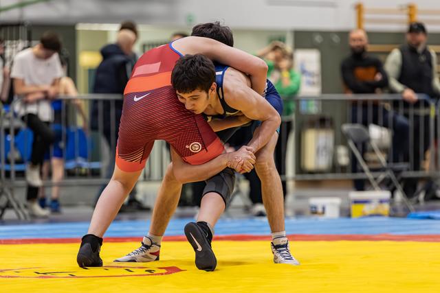 Young wrestler in red singlet locks hands around opponent in blue, both bent low in intense grapple on yellow mat.