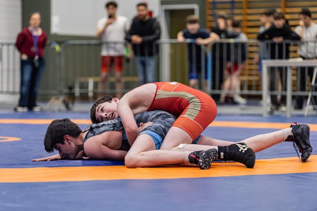 Young wrestler in red singlet dominates opponent, pinning him face-down on the orange and blue mat.