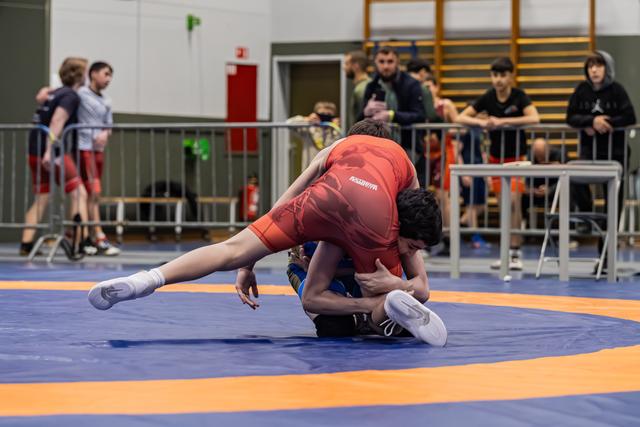Wrestler in red singlet executes a takedown, pinning opponent to the blue mat as spectators watch from behind barriers.