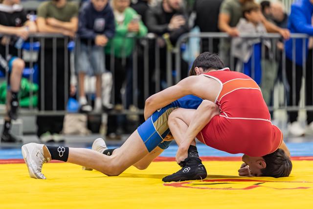 Wrestler in red singlet drives opponent in blue to the mat, executing a powerful takedown during a competitive bout.