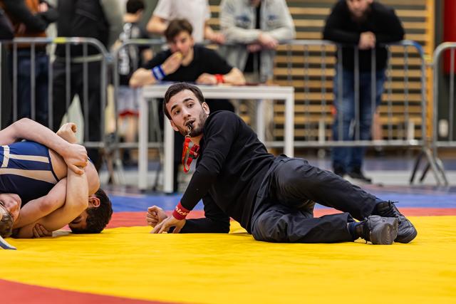Referee lying on yellow mat blows whistle while closely watching two wrestlers grapple during indoor tournament.