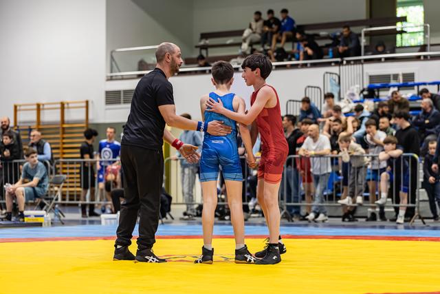 Coach and referee shake hands beside two young wrestlers in blue and red singlets after a bout on the mat.