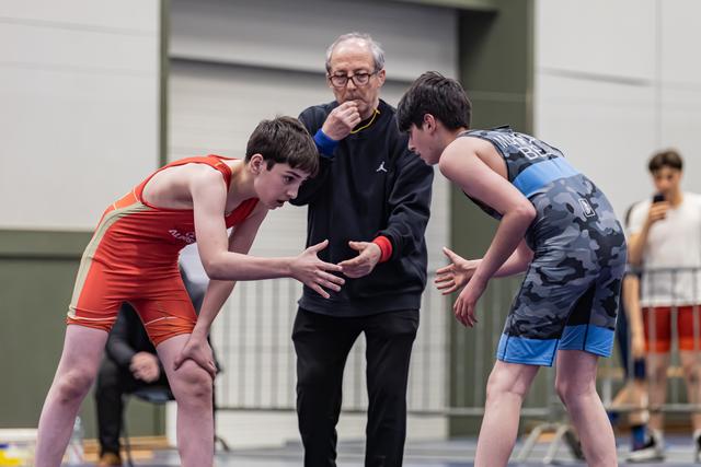 Two young wrestlers lean in to shake hands before a bout, overseen by a referee blowing a whistle indoors.