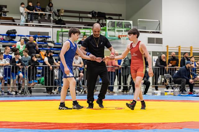 Referee joins hands of two young wrestlers in blue and red singlets before their bout on a colorful mat.
