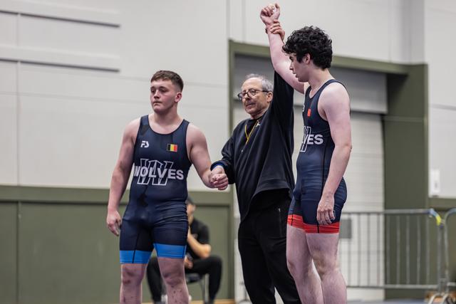 Referee raises the fist of a victorious wrestler in a navy Wolves singlet, while his defeated opponent stands nearby.