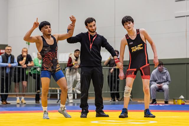 Referee raises the left arm of a jubilant wrestler in tiger singlet as the defeated opponent stands solemnly beside them.