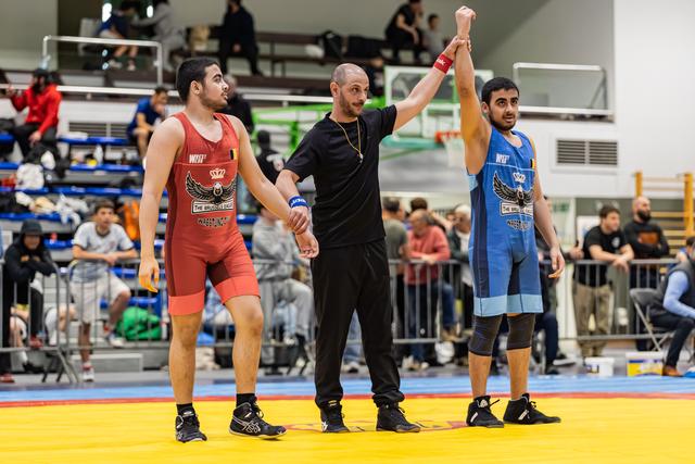 Referee raises blue-singlet wrestler's arm in victory while red-singlet opponent stands dejected on yellow mat.