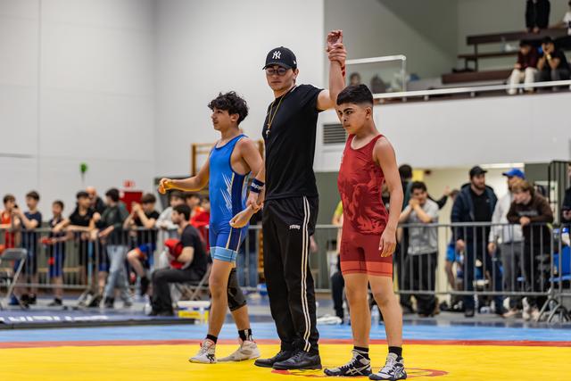 Referee raises the arm of a young wrestler in red singlet declaring victory, while defeated opponent in blue walks away.