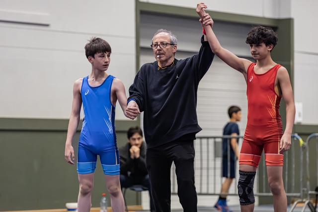 Referee raises the arm of the red-suited winner while the blue-suited boy stands stoic beside them in a gym.