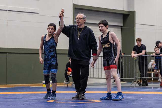 Referee raises the arm of a young wrestler in blue singlet declaring victory, as the opponent stands beside them.