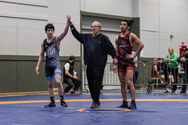 Referee raises the left wrestler's hand in victory as the opponent stands aside, looking composed on the blue mat.