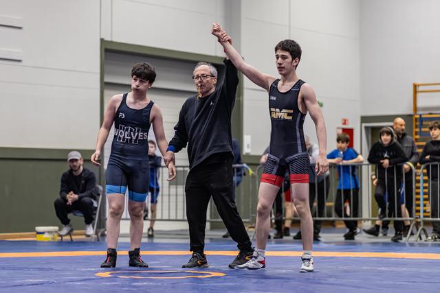 Referee raises the arm of the victorious wrestler in a black singlet, while the defeated opponent stands beside them.