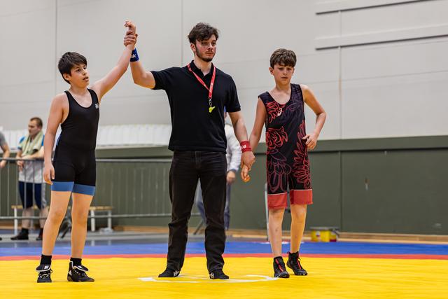 Referee raises young wrestler's hand in victory while defeated opponent stands dejected on yellow mat.