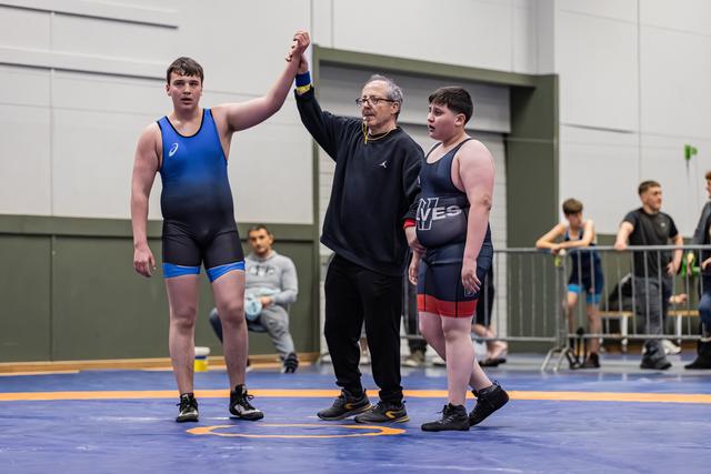 Referee raises the hand of a young wrestler in blue singlet declaring victory, as opponent stands nearby on blue mat.