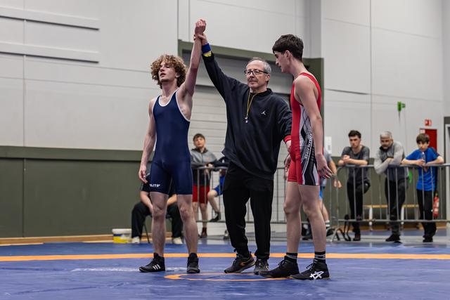 Referee raises the red-singlet wrestler's hand in victory while the blue-singlet opponent looks away, dejected.