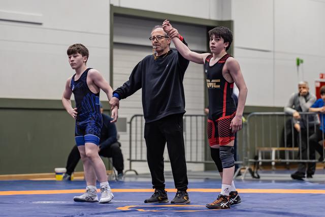 Referee raises the arm of a young wrestler in black-red singlet, declaring victory on a blue mat indoors.