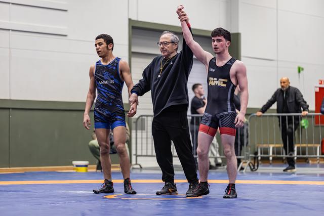 Referee raises the right arm of a victorious wrestler in black singlet, while defeated opponent in blue looks down.