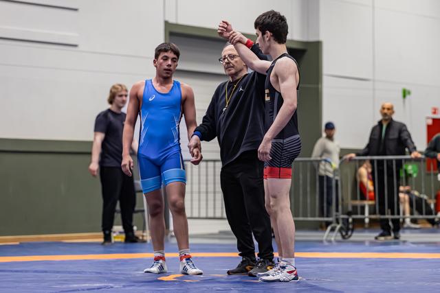 Referee raises the arm of a young wrestler in black, declaring victory, while the defeated opponent in blue stands nearby.