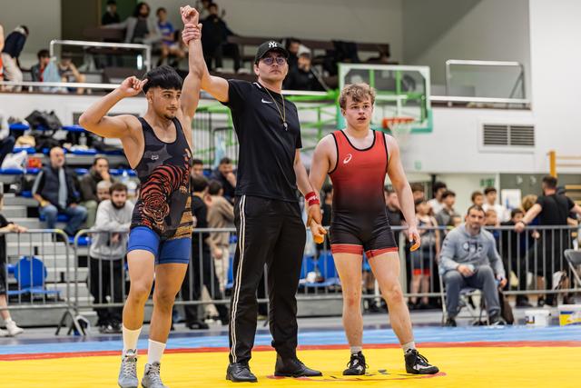 Referee raises the arm of a victorious wrestler in a tiger singlet; the defeated opponent stands quietly beside them.