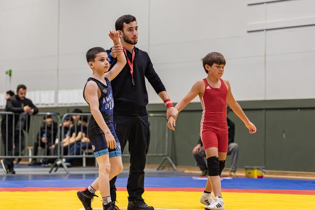 Referee raises the arm of a young wrestler in blue singlet declaring victory, as defeated opponent in red walks away.