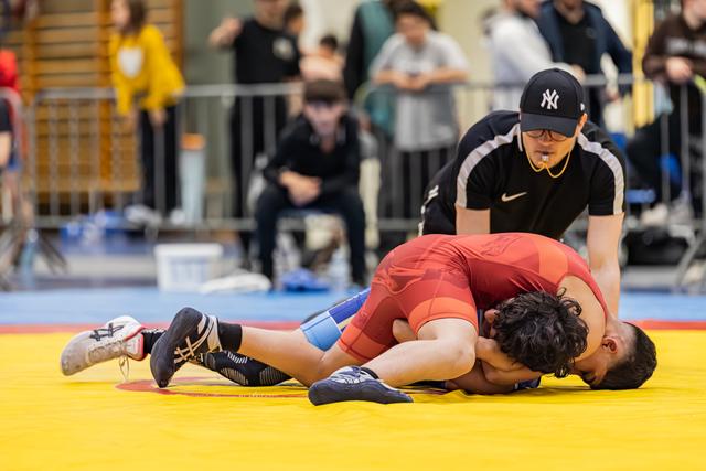 Referee in NY cap watches closely as wrestler in red singlet pins opponent face-down on yellow mat.
