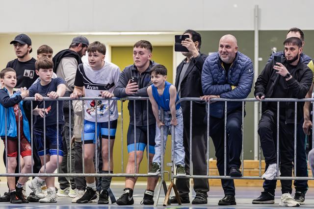 Coaches, athletes, and parents lean on metal barriers, watching intently with phones raised, at an indoor wrestling event.