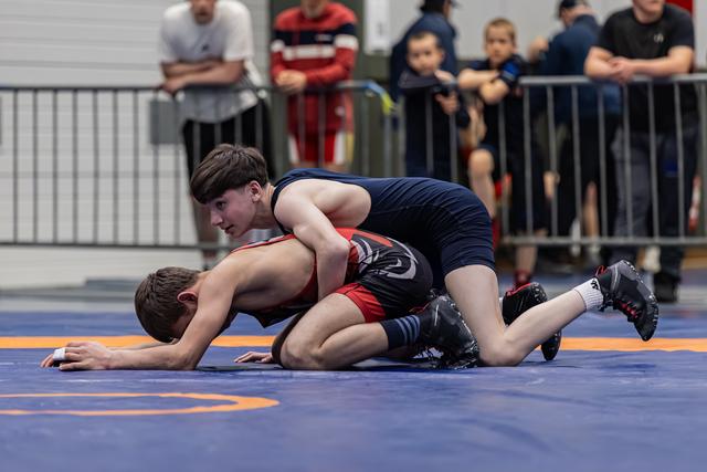 Young wrestler in navy singlet controls opponent on the mat, focused expression, spectators watching in background.