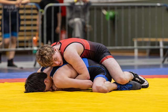 Teenage wrestler in red singlet pins dark-haired opponent to yellow mat, applying controlled pressure during match.